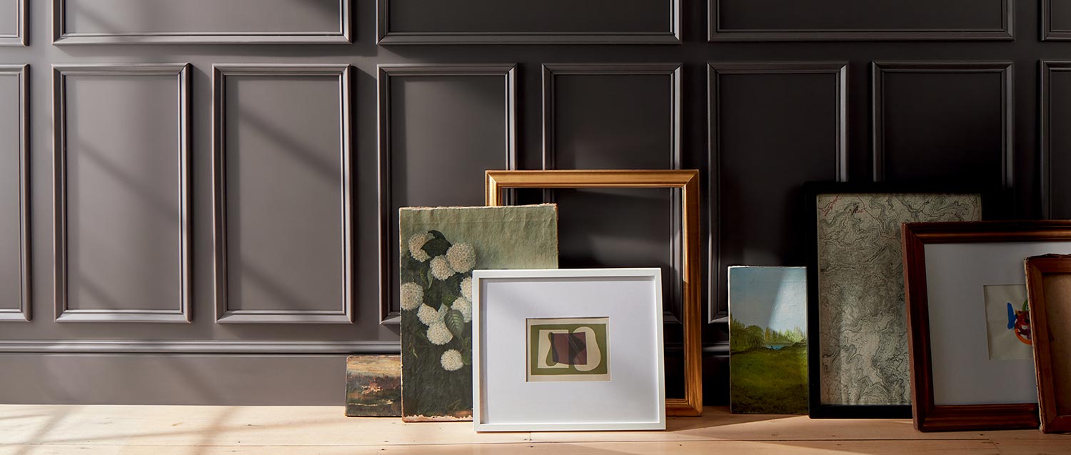 Artwork and frames arranged on a floor against a deep chocolate brown-painted wall with decorative moulding.